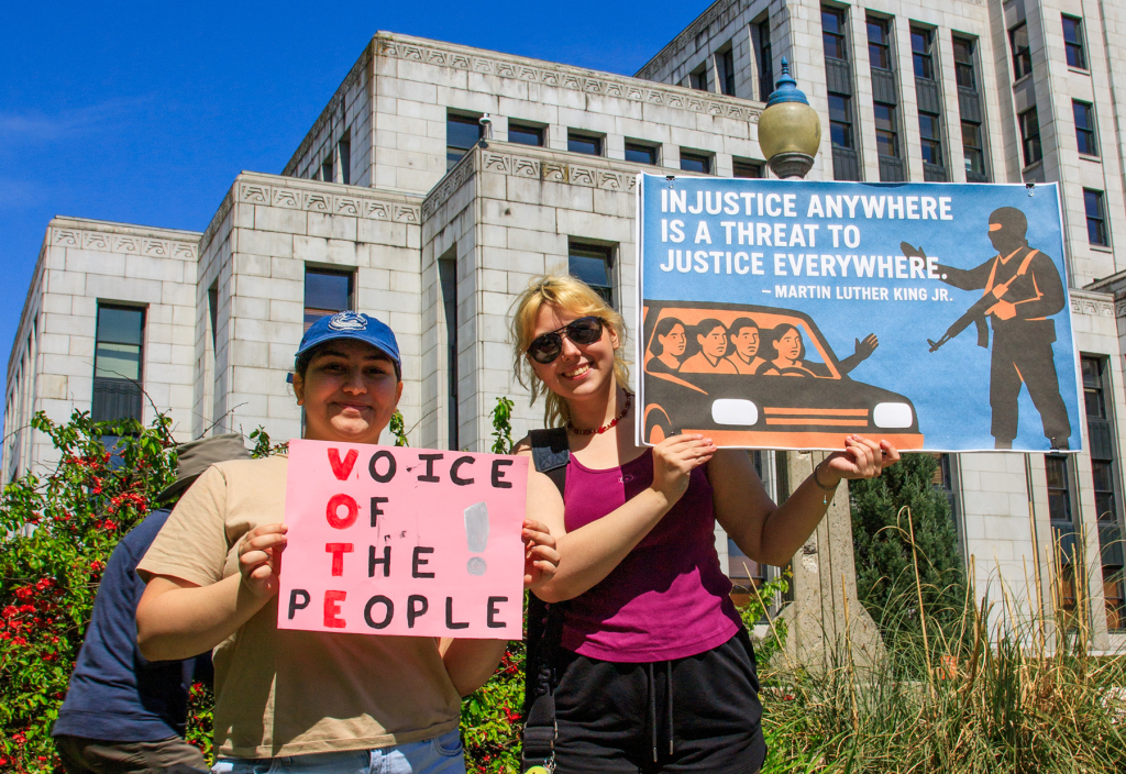Two young women, smiling, in the sun in front of the art deco Vancouver City Hall, holding protest signs against authoritarianism.