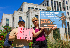 Two young women, smiling, in the sun in front of the art deco Vancouver City Hall, holding protest signs against authoritarianism.